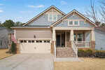 A modern suburban house with gray siding, stone accents, a two-car garage, a front porch with steps, and an American flag by the driveway. The yard has brown grass and light landscaping.