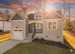 A modern gray house with white trim, a single-car garage, front porch, and stone accents sits under a vibrant sunset sky, surrounded by trees and a well-kept lawn.