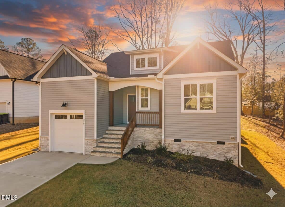 A modern gray house with white trim, a single-car garage, front porch, and stone accents sits under a vibrant sunset sky, surrounded by trees and a well-kept lawn.