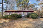 A small brick house with red trim, a central front door, large bushes in front, and tall trees in the background, photographed on a sunny day.