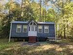 A small blue cabin with white trim sits on a grassy clearing surrounded by tall trees. The cabin has a metal roof, three front windows, a white double door, steps with red paint, and a picnic table on the right.