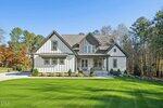A modern farmhouse-style home with white vertical siding, black trim, and a front porch, set on a well-manicured lawn with trees on either side under a clear blue sky.