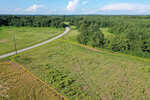 Aerial view of a grassy, vacant plot of land bordered by a yellow outline, next to a curving rural road and surrounded by green trees and fields under a partly cloudy sky.