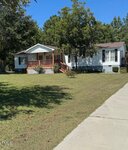 A white, single-story house with a covered front porch and brown railings sits on a large, grassy yard bordered by trees, next to a paved walkway.