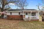 A small, single-story house with red brick on the lower half and light-colored siding on top. It has a covered front porch with railings, steps, and a large tree in the yard. The grass is patchy and some leaves are on the ground.