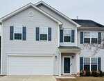 Two-story suburban house with light gray siding, blue shutters, and a double garage. There are several windows on both floors, a small front porch with a black door, and a neatly trimmed lawn with small shrubs.