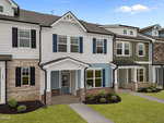 A row of modern townhouses with brick and siding exteriors, each featuring a small front porch, manicured lawns, and neatly trimmed shrubs under a bright blue sky.