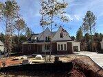 A two-story modern house under construction with red brick and white siding, a covered front porch, and a sloped driveway. Tall trees surround the property, and construction materials are scattered in the yard.