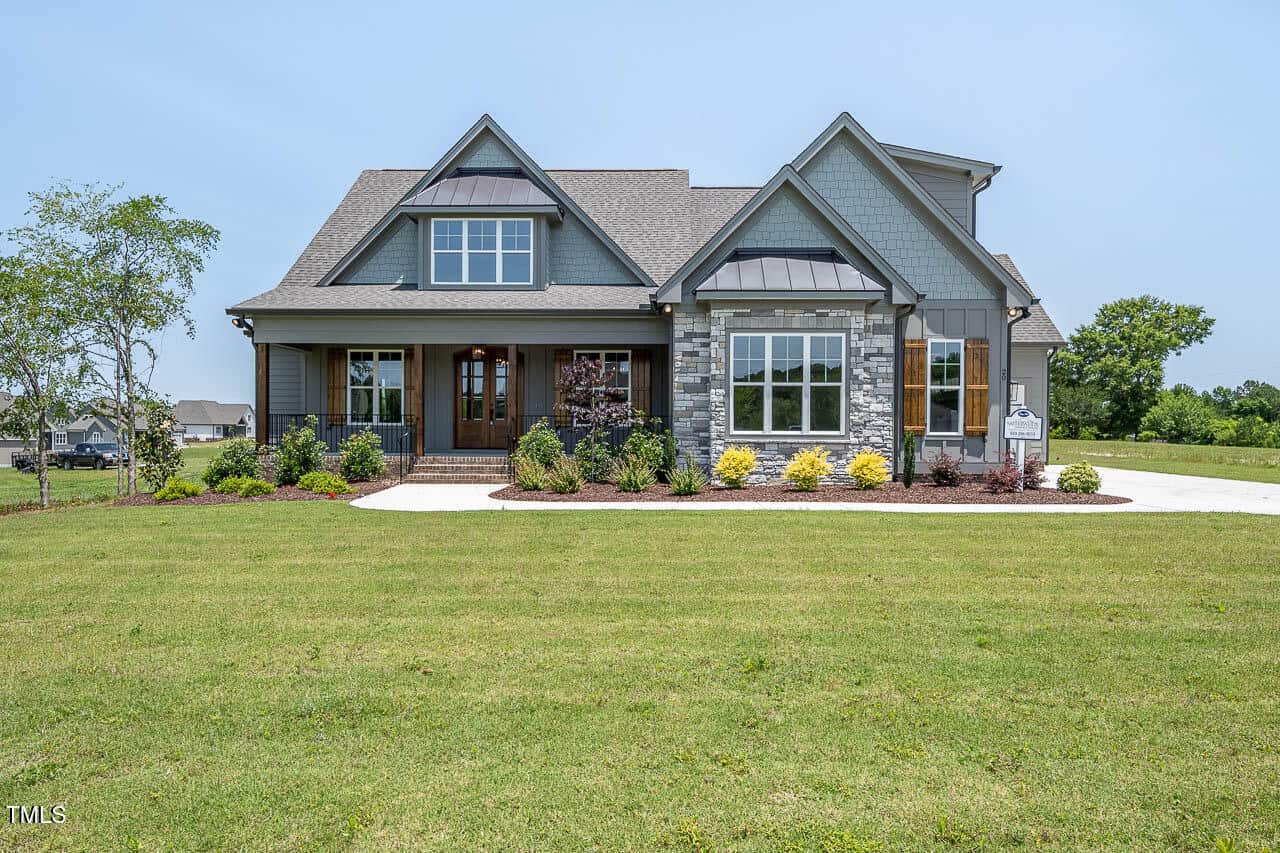 A modern two-story house with gray siding, stone accents, a covered front porch, and landscaped yard sits on a spacious green lawn under a clear blue sky.