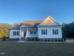 A single-story white house with a front porch, gable roof, and brick foundation sits on a grassy lawn, bordered by trees under a clear blue sky.