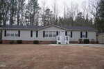 A single-story gray manufactured home with white trim, black shutters, and a small front porch sits on a large, brown grass lawn, backed by leafless trees and surrounded by woods under a cloudy sky.
