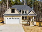 A modern two-story beige house with white trim, a covered front porch, and a double garage. The house is surrounded by a forest and has a paved driveway and small landscaped area in front.