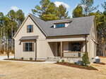 A modern beige house with a gable roof, front porch, and wooden shutters, surrounded by a dry lawn and pine trees in the background under a clear sky.