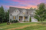 Gray two-story house with front porch, gabled roof, and attached white garage, surrounded by green lawn and trees, with a vibrant pink and purple sunset sky in the background.