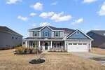 Two-story suburban house with gray siding, blue gable accents, white trim, and a covered front porch. A two-car garage is on the right, and the yard is landscaped with small trees and shrubs under a partly cloudy sky.