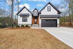 Modern two-story house with white board-and-batten siding, black trim, a double garage, stone accents, and a concrete driveway. The yard is mostly bare with a few shrubs near the entry. Trees and neighboring homes are visible.