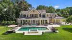 A large, beige two-story house with dormer windows, a covered back porch, and an in-ground rectangular pool with a hot tub, surrounded by green lawn, trees, and outdoor seating areas.