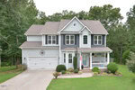 Two-story suburban house with gray siding, black shutters, and a covered front porch. The home has a double garage, manicured lawn, hedges, a small American flag, and is surrounded by trees.
