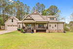 A large modern farmhouse with beige siding, stone accents, a covered front porch, and an attached two-car garage, set on a spacious, grassy lawn bordered by tall pine trees.