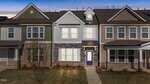 A two-story townhouse with a mix of white, tan, and sage green paneling, dark stone accents, white trim, and a blue front door. Small shrubs and a young tree are planted in the front yard at dusk.