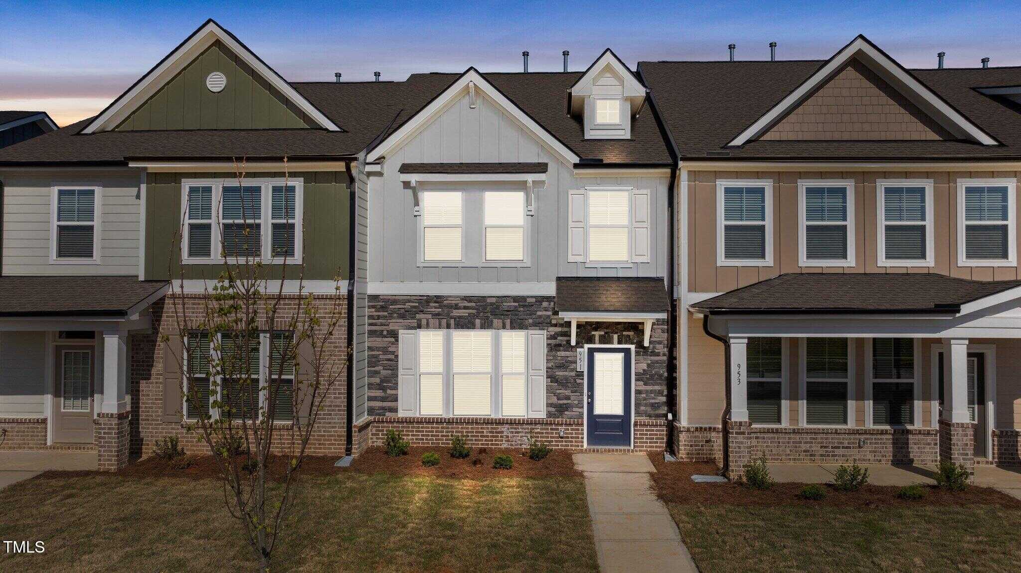 A two-story townhouse with a mix of white, tan, and sage green paneling, dark stone accents, white trim, and a blue front door. Small shrubs and a young tree are planted in the front yard at dusk.
