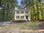 A two-story yellow house with black shutters, white trim, and a covered front door, surrounded by tall trees and greenery, with a gravel driveway to the left.