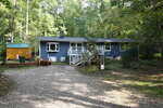 A blue single-story house with white trim sits in a wooded area. A gravel driveway leads to the front steps, and a small yellow shed is on the left side. Trees and greenery surround the house.