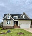 A modern two-story house with gray siding, brick accents, and a black roof. It has a double garage, wooden front door, and newly planted grass and landscape features in the front yard under a cloudy sky.