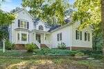 Two-story white house with dark shutters, a front porch, and bay windows surrounded by greenery and trees. Brick foundation and a landscaped yard with stone-edged flowerbeds in front.