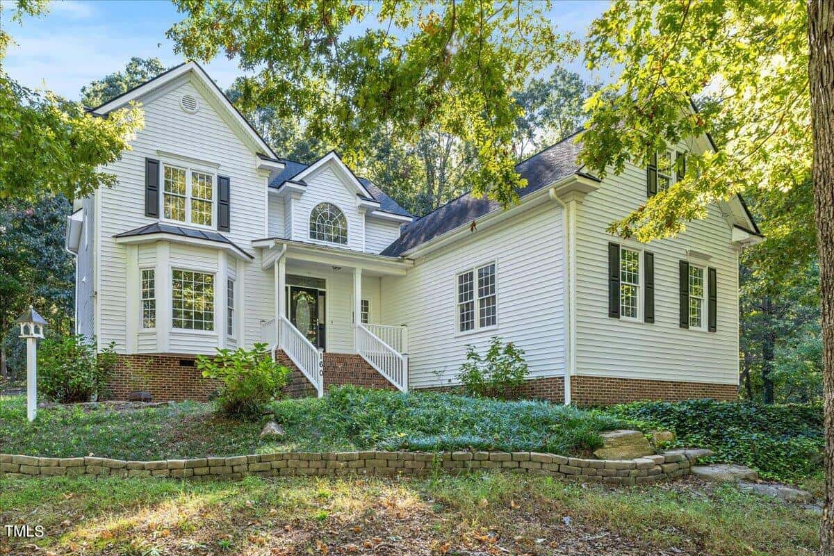 Two-story white house with dark shutters, a front porch, and bay windows surrounded by greenery and trees. Brick foundation and a landscaped yard with stone-edged flowerbeds in front.