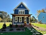 A two-story blue house with white trim and a covered front porch sits on a landscaped lawn with flower beds and pumpkins by the entrance, under a clear sunny sky.