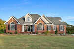 A large, two-story brick house with white trim, multiple arched windows, a gray shingled roof, and a manicured lawn, set against a blue sky and trees in the background.