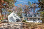 A two-story house with a large front porch and attached garage sits among tall autumn trees. A driveway leads to the garage, with a parked truck and a boat visible on the left side.
