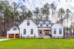 A modern white farmhouse with black trim, wood accents, and a large front porch. The house has a gable roof, dormer windows, a garage with a wooden door, and is surrounded by tall pine trees and a green lawn.