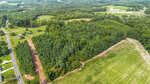 Aerial view of a rural landscape with a forested area, cleared land, open fields, a road, scattered houses, and a small pond surrounded by greenery. Construction equipment is visible near the trees.