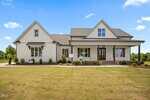 A modern farmhouse-style home with white siding, black trim, and a covered front porch, set against a bright blue sky with lush greenery in the background and a neatly trimmed lawn in front.