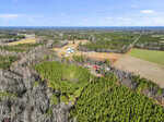 Aerial view of a rural landscape with green forests, open fields, and scattered buildings under a blue sky. A cluster of farm structures and a red-roofed building are visible near the center of the image.