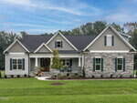 A modern suburban single-story house with gray siding, stone accents, gabled roof, and a wooden front door. Manicured lawn, small shrubs, and a young tree decorate the front yard under a partly cloudy sky.