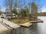 A small house with a porch sits near a wooded shoreline, next to a curved wooden dock extending into a calm lake. Leafless trees surround the property, and the sky is partly cloudy.
