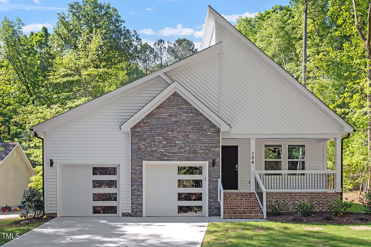 Modern white house with a stone accent facade, two garage doors, a covered front porch with railings, and steps leading to the entrance. Surrounded by green trees on a sunny day.