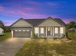 Single-story suburban house with a two-car garage, front porch, and neatly landscaped yard at dusk under a vibrant purple and pink sky. The house has gray siding and stone accents.