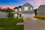 A modern two-story house with blue-gray siding, white trim, a double garage, a covered front porch, and a well-manicured yard. The driveway leads up to the house, and the sky is colorful at sunset.