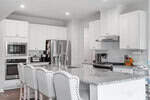 Bright, modern kitchen with white cabinets, stainless steel appliances, granite countertops, and a large island with four upholstered chairs. Kitchen tools and small appliances are visible on the counters.