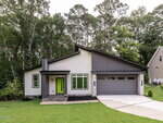 Modern single-story house with gray siding, stone accents, and a sloped roof. It features a bright green front door, double garage, manicured lawn, and is surrounded by tall trees and greenery.