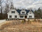 A modern farmhouse with white siding, stone accents, black roof, and multiple gables, set in front of a wooded area with a grassy field and a paved driveway leading to the entrance.