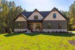A modern two-story house with gray siding, stone accents, gabled roof, and large windows, set amid green grass and surrounded by tall trees on a sunny day. A small front porch has two rocking chairs.