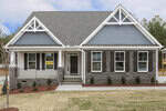 A newly built single-story house with gray siding, stone accents, and white trim. The home has a covered front porch, dark shutters, a gray shingle roof, and a freshly landscaped yard with small shrubs.