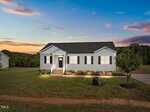 A single-story gray house with white trim and black shutters sits on a grassy lawn at sunset, with a front porch, landscaped garden, and a paved driveway. Trees and fields are visible in the background under a colorful sky.