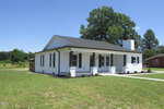 A single-story white brick house with a black roof, a front porch supported by white columns, and large windows, surrounded by grass and a few small shrubs, under a clear blue sky.