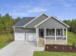 A modern single-story gray house with a covered front porch, white trim, and a double garage. The house sits on a neatly landscaped yard with green grass, shrubs, and a concrete driveway, surrounded by trees.